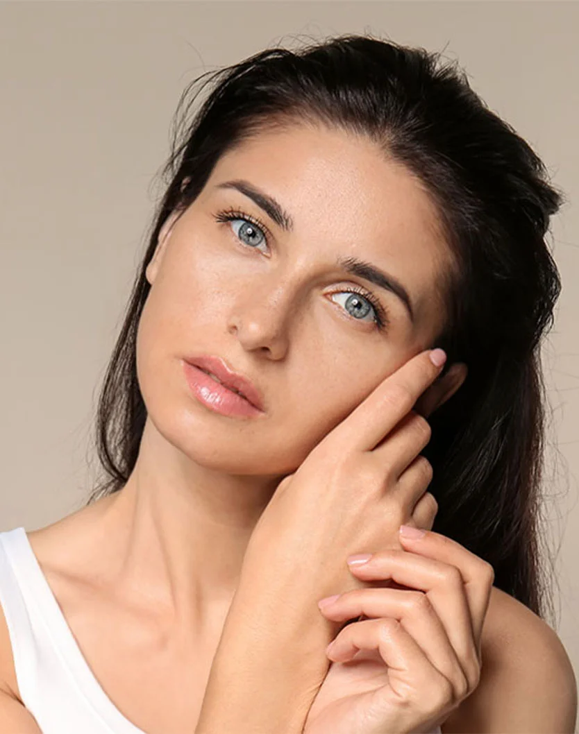 A woman with long dark hair and striking blue eyes gently touches her face, set against a soft neutral background. - Acne Scar Treatment in New York, NY
