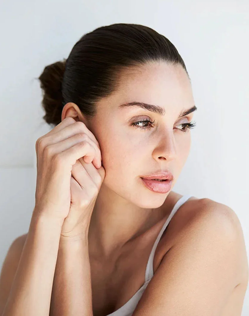 A woman gazes thoughtfully, resting her chin on her hand, with a natural look and minimal makeup against a light background. - Brow Lift in New York, NY
