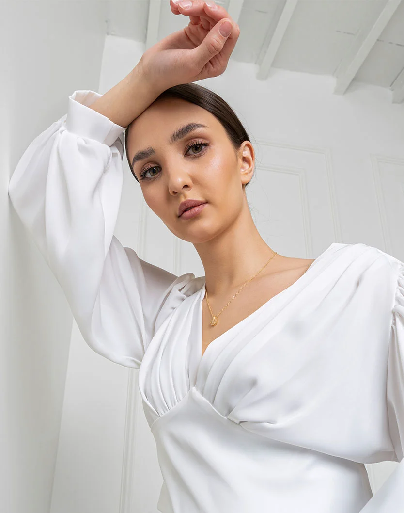 A woman in a white blouse gazes thoughtfully at the camera, her hand raised above her head against a light background. - Droopy Eyelid Repair in New York, NY