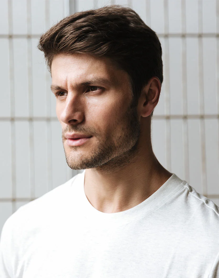 A close-up of a young man with short, styled hair, wearing a plain white t-shirt, gazing thoughtfully to the side. - Earlobe Reduction in New York, NY