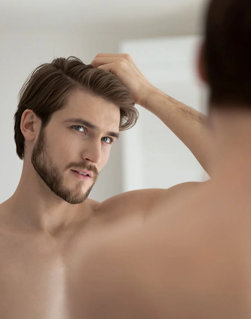 A young man with a beard styles his hair while looking at his reflection in a mirror, showcasing a thoughtful expression. - Hairline Lowering in New York, NY