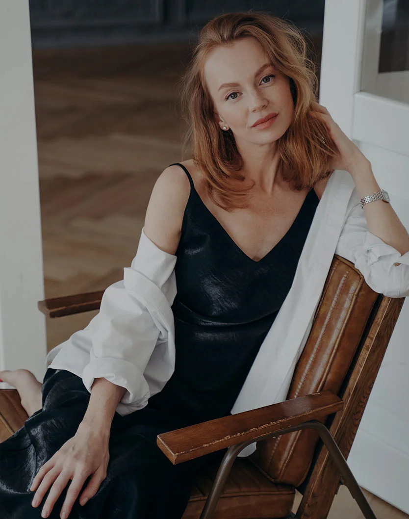 A woman with wavy hair sits casually in a wooden chair, wearing a black dress and a white oversized shirt, exuding elegance. - Laser Skin Resurfacing in New York, NY