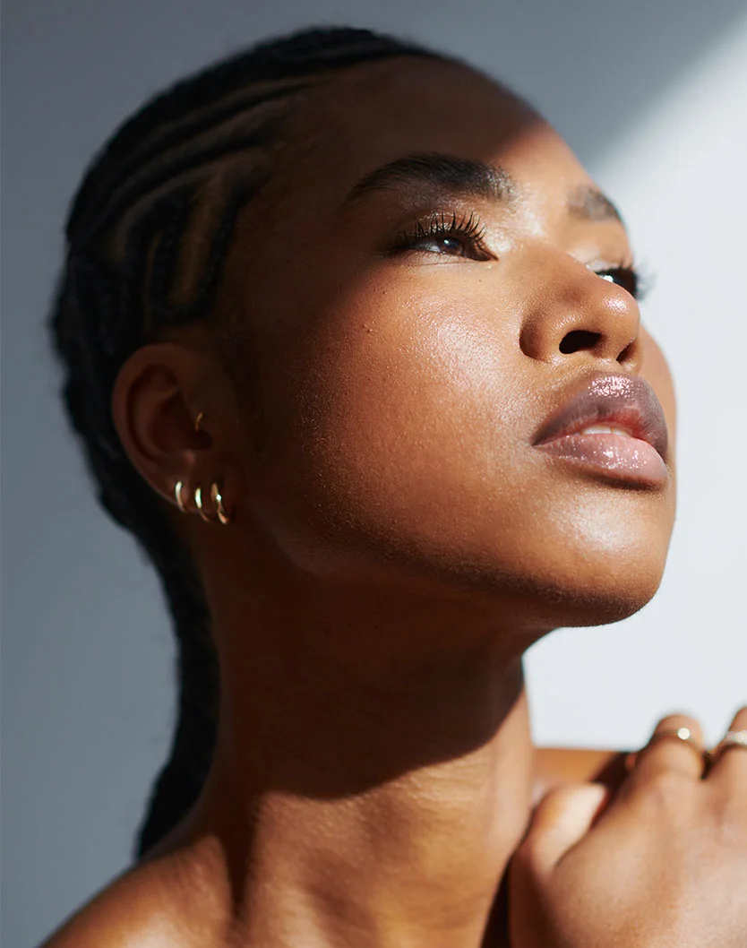 A young woman with braided hair gazes upward, illuminated by soft natural light, showcasing her radiant skin and elegant earrings. - Lip Reduction in New York, NY