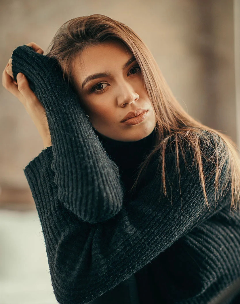 A young woman with long brown hair rests her face on her arm, wearing a black sweater in a softly lit indoor space. - Lower Eyelid Surgery in New York, NY