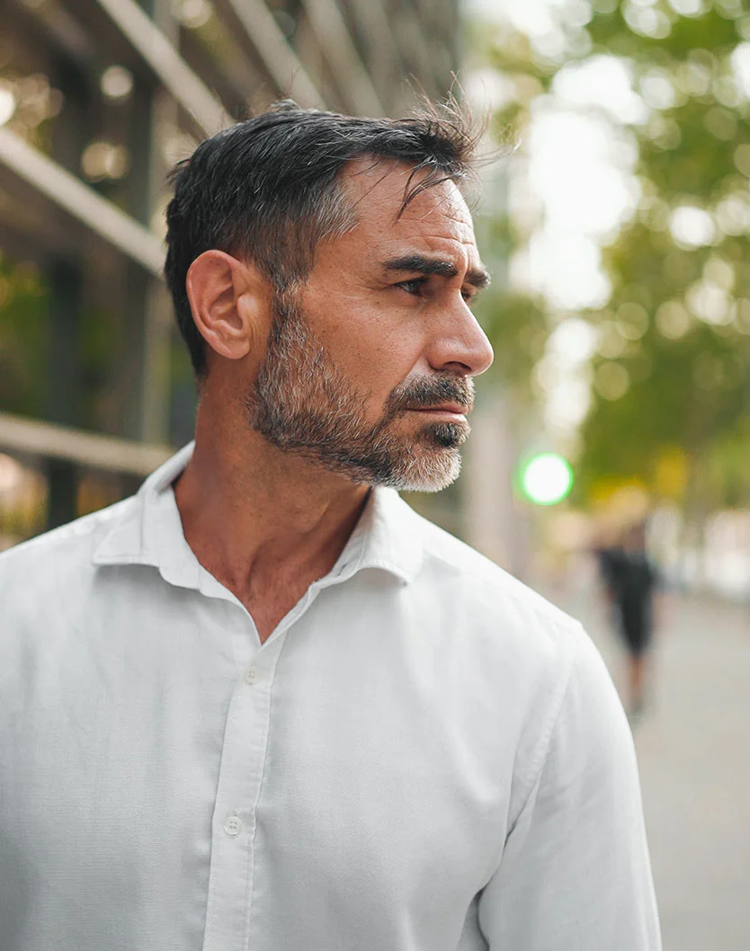 A shot of a side profile of a thoughtful man with a salt-and-pepper beard stands outdoors, gazing into the distance, with blurred greenery behind him. - Rhinophyma Treatment in New York, NY