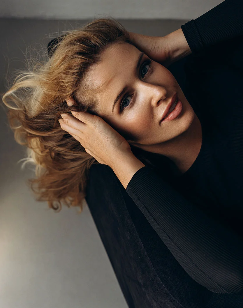 A slightly high-angle close-up of a woman with tousled, shoulder-length light brown curls. Her hands rest on her head, and she gazes slightly upward with a relaxed, thoughtful expression. She has fair, smooth skin and wears a dark, ribbed long-sleeved top. The softly lit, muted gray background is out of focus, emphasizing her features. - Tear through Filler in New York, NY