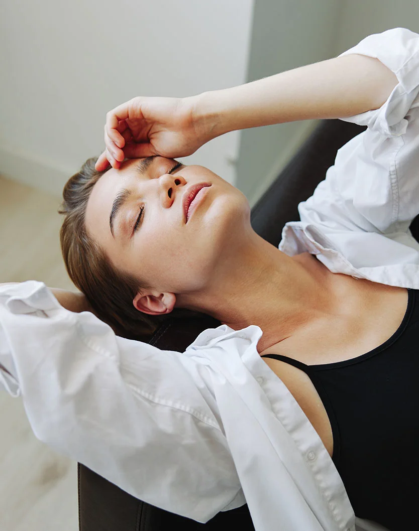 A young woman reclines, her head resting in her hand with eyes closed and a serene expression. She wears a white long-sleeved button-down shirt over a black tank top. Her light brown, tousled hair falls around her shoulders, and her smooth, light-toned skin is softly lit. The background features a light neutral wall, with a dark-colored surface behind and under her, creating a calm and relaxed atmosphere. The lighting is soft and even, without harsh shadows. - Upper Eyelid Surgery in New York, NY